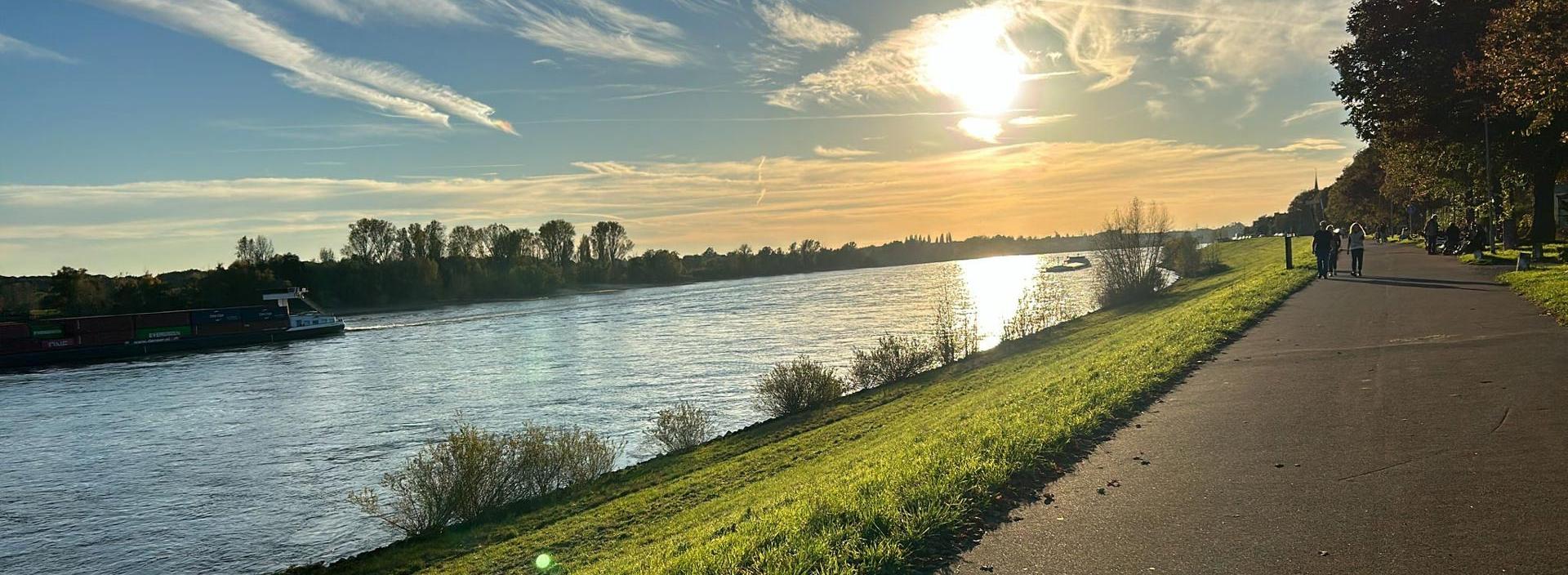 Man sieht den Rhein und am Horizont den blauen Himmel mit Kondenzstreifen und der untergehenden Sonne