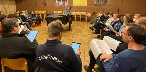 Sie sehen die Sängerinnen und Sänger des Chores Caminando im Stuhlkreid singen, in der Mitte steht ein Flügel